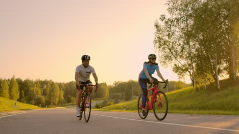 Dos-Ciclistas,-Un-Hombre-Y-Una-Mujer,-Viajan-Por-La-Carretera-En-Bicicletas-De-Carretera-Con-Cascos-Y-Ropa-Deportiva-Al-Atardecer-En-Cámara-Lenta